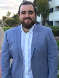 author photo: dg lewis with combed back hair, full beard, wearing a blue and pink plaid suit jacket over a white button-down shirt